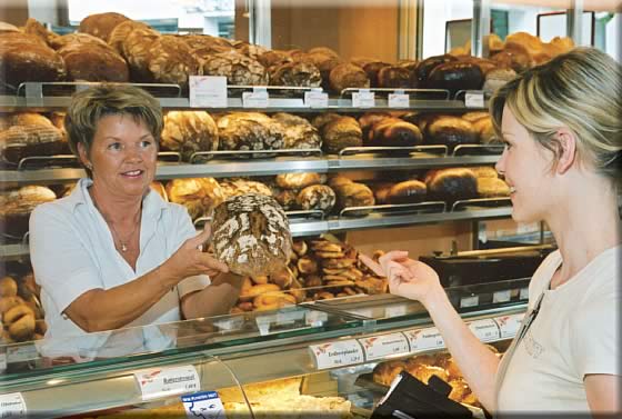 Eine Verkäuferin reicht einer jungen Kundin in einer Bäckerei ein Brot, im Hintergrund ein mit Broten gefülltes Brotregal 